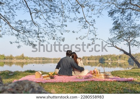Young couple in love sitting on blanket with picnic basket and embracing to watching lake view. Royalty-Free Stock Photo #2280558983