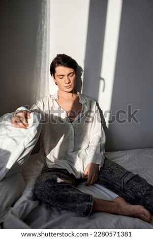 Handsome man in pajamas with closed eyes in white shirt and trousers sits on bed at home, close up photo. good morning.