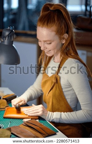 smiling ginger girl enjoys burnishing edges of leather.closeup side view photo, craftswoman working, using edge slicker,necessary tools for tailor,accurate process of sewing,making wallet Royalty-Free Stock Photo #2280571413