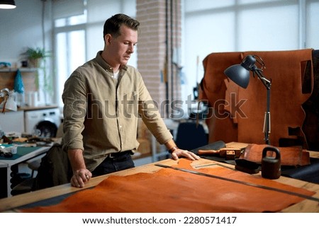 handsome man has bought genuine leather, he is standing behind the table, loooking at fabric and thinking how to sew. close up side view shot, free time, lifestyle, hobby. business
