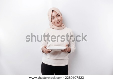 A smiling Asian Muslim woman is fasting and hungry and holding and pointing to a plate 