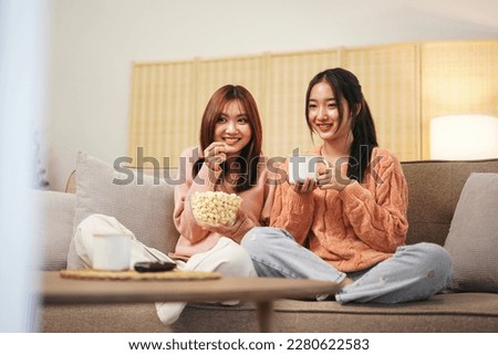Young woman lesbian couple in sweater sitting on the comfortable sofa to watching cinema together in living room at home and eating delicious popcorn with drinking hot chocolate.