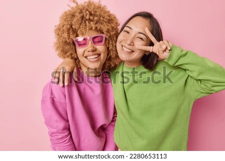 Horizontal shot of positive women friends smile and embrace make peace gesture have happy expressions dressed casually isolated over pink background. People and emotions concept