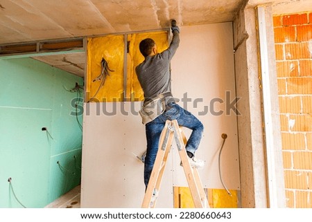 Worker on wooden ladder holds professional manual power tool ACU drill and tightens the screws on plasterboards installed on the partition dry wall insulated with mineral wool. Royalty-Free Stock Photo #2280660643