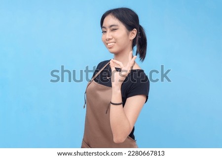 A young and confident asian female waitress or barista winks while making the ok sign. Wearing a brown apron and black shirt welcoming a customer and against a light blue background.