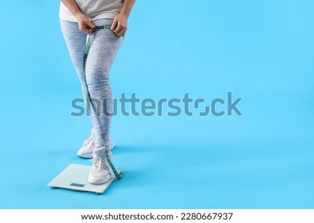 Young overweight woman with scales measuring her leg on blue background Royalty-Free Stock Photo #2280667937