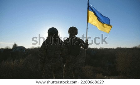Female and male soldier of ukrainian army standing at peak of hill with raised flag of Ukraine. Military couple in camouflage uniform holds hands of each other looking at blue sky with sun. 