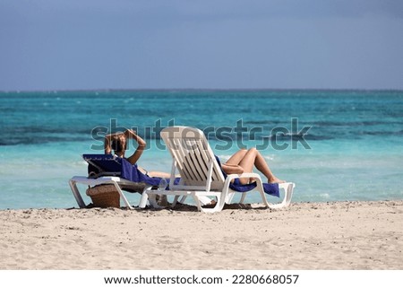 Two girls in bikini tanning on deck chairs on sea waves background. Sandy beach on Atlantic ocean coast