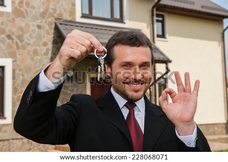 closeup on smiling real estate agent ready to sell house. Male real etate agent in front of home holding keys and ok sign