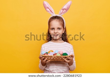 Portrait of smiling little girl with rabbit ears on her head with basket of colored eggs in her hands posing isolated on yellow background, celebrating holiday.
