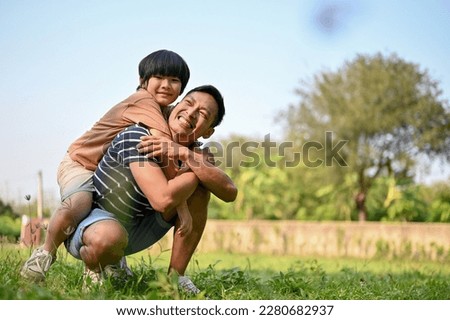 Smiling and happy Asian dad playing with his son in the backyard while carrying him on his back. piggyback pose. happy family and single dad concept Royalty-Free Stock Photo #2280682937