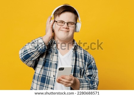 Young satisfied man with down syndrome wear glasses casual clothes headphones listen to music use mobile cell phone isolated on pastel plain yellow color background. Genetic disease world day concept Royalty-Free Stock Photo #2280688571