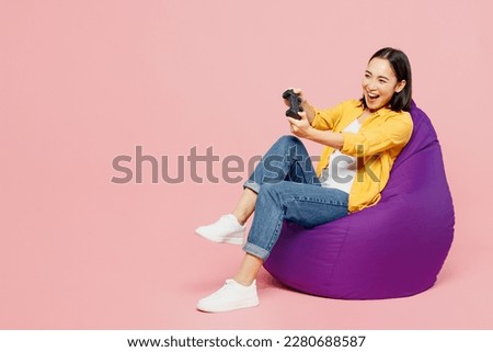 Full body young woman of Asian ethnicity wear yellow shirt white t-shirt sit in bag chair hold in hand play pc game with joystick console isolated on plain pastel light pink background studio portrait
