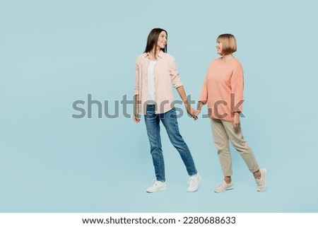 Full body side view fun elder parent mom with young adult daughter two women together wearing casual clothes hold hands walk go look to each other isolated on plain blue background. Family day concept