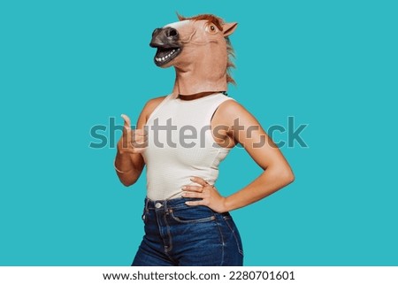 Young funny woman in a horse comical mask, does thumbs up sign with hand at studio, isolated over blue background. Carnival, humorous concept. Royalty-Free Stock Photo #2280701601