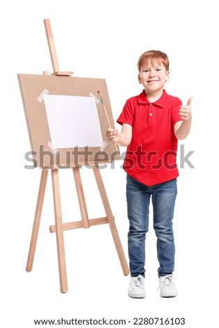 Little boy with brush near easel with canvas showing thumbs up against white background. Creative hobby