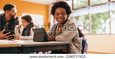 Boy smiling at the camera while sitting with a digital tablet in a classroom. Primary school kid reads an ebook at school. Young child developing his reading skills using modern technology. Royalty-Free Stock Photo #2280717423