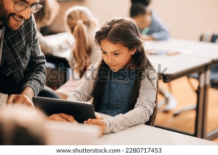 Teacher shows a student how to use a digital tablet in an elementary school. Male tutor teaches digital literacy and coding to young children. Child education and mentorship using modern technology. Royalty-Free Stock Photo #2280717453