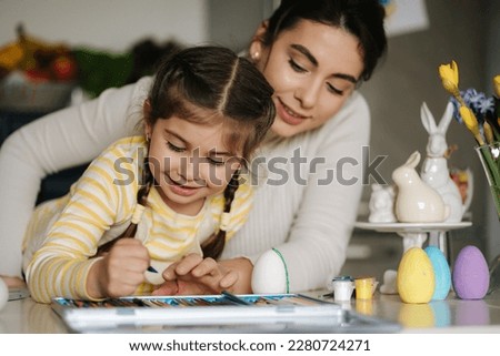 Happy laughing little girl painting picture with mom at the table in the kitchen. Easter holiday. Spring time