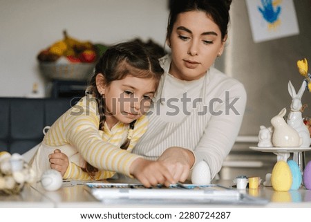 Mom and her cute little girl sitting at the table in the kitchen and color the picture with pencils
