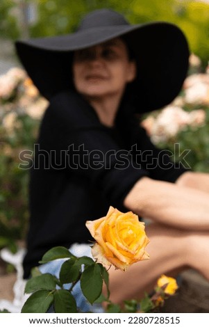 Blurred image of a seated woman in a large black hat, looking away, in the background and a large blooming yellow rose in the foreground, selective focus.