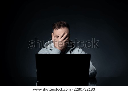 Man in a white hoodie sitting at a table in front of a laptop makes a facepalm on a black background.