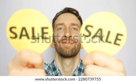 Close-up of a cute smiling man showing two defocused yellow round signs with the word SALE