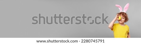 Happy boy with bunny ears holding Easter egg near eye on light grey background. Banner design