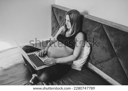 Pregnant student girl studying at home, learning webinar, workshop. Woman writing notes and using a laptop in bedroom. Female have computer as freelancer. Remote work, overwork. Black and white photo.