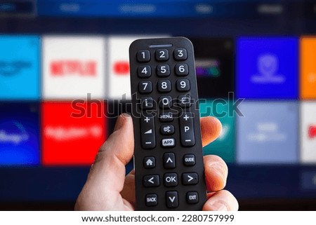 A man is holding the remote control of a smart TV with a television screen in the background with some blurry video streaming service app icons. Man using a tv remote control. Royalty-Free Stock Photo #2280757999