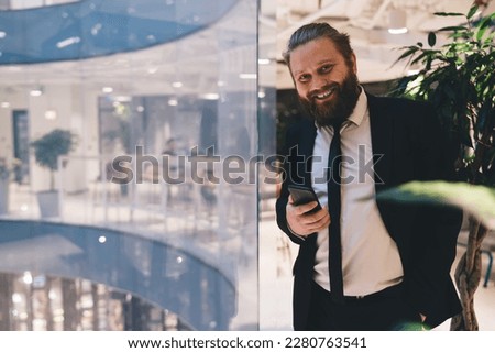 Positive male entrepreneur in suit smiling and looking at camera while standing in hallway of modern office with hand in pocket and using cellphone during workday
