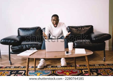 Shocked African American male freelancer showing hands at monitor while looking at screen of laptop and sitting at wooden table with smartphone cup of coffee eyeglasses in living room at home