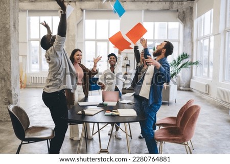 Cheerful multiracial coworkers standing together around table while raising hands up and tossing colorful papers in air at workplace during success celebration Royalty-Free Stock Photo #2280764881
