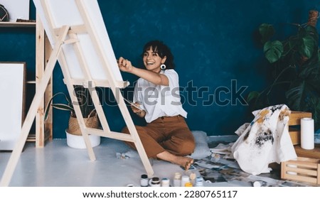 Excited young female artist draws  picture with paints and enjoys relaxing process of her hobby.  Creative happy brunette woman creates picture on easel in studio. Learning to draw with oil