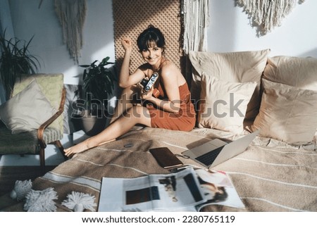 Full body of cheerful Hispanic photographer with vintage camera sitting on bed and smiling while working on laptop in light bedroom and looking at camera