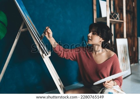 Attractive young female artist draws  picture with paints and enjoys relaxing process of her hobby.  Creative and talented brunette woman creates picture on easel in studio. Learning to draw with oil