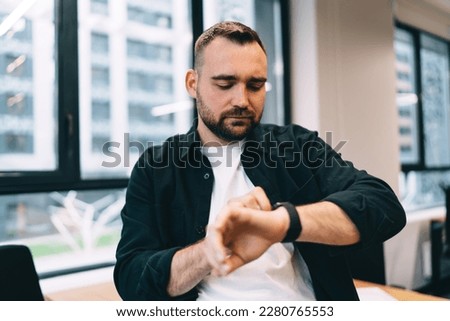 Bearded man in casual clothing looking at wristwatch while waiting for end of workday hurrying to meeting standing in office Royalty-Free Stock Photo #2280765553