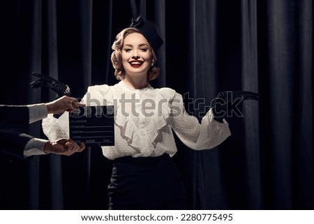 Film shooting. Charming actress wearing vintage clothes and posing at camera and smiling over dark background. Concept of old films, actress, beauty, fashion, old films, actress, retro, vintage Royalty-Free Stock Photo #2280775495