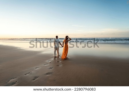 Romantic couple walking on sunset beach, enjoying evening light, relaxing on tropical summer vacation. Honeymoon. Love. Back view. Woman wearing orange maxi dress, man in linen pant and white shirt.
 Royalty-Free Stock Photo #2280796083
