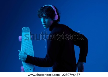 Male smiling teenager in headphones listening to music with skateboard in hand, portrait dark blue background, neon light, style and trends, mixed light, men's fashion, copy space