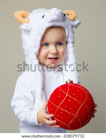 Portrait of Caucasian baby with blue eyes wearing a sheep hat with yellow horns and white shirt standing on a light background holding red golden ornament decoration, New Year concept, studio