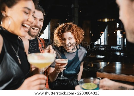 Carefree redhead woman spending time with interracial friends holding cocktails in bar
