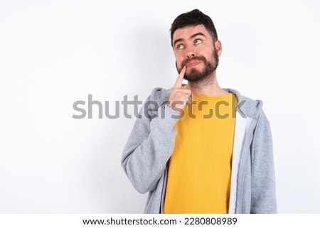 Lovely dreamy Young caucasian mán wearing trendy clothes over white background keeps finger near lips looks aside copy space. Royalty-Free Stock Photo #2280808989