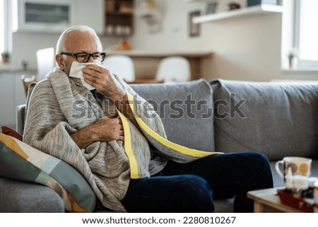 A senior Caucasian man is lying in bed with a bad cold and is blowing his nose using a napkin. An older ill man with temperature is lying in his bed covered with a blanket.  Royalty-Free Stock Photo #2280810267