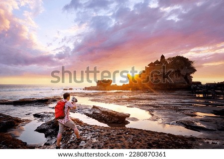 Travel and photography. Young man with camera taking picture of beautiful balinese landscape. Ancient hinduism temple Tanah lot on the rock against sunset sky. Bali Island, Indonesia.