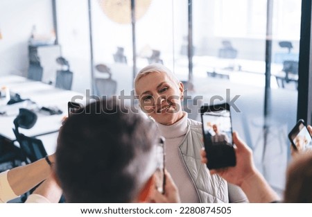 Smiling senior female with shorthair looking at camera while standing and giving pose to unrecognizable colleagues to shoot pictures in smartphones during daytime in blurred office