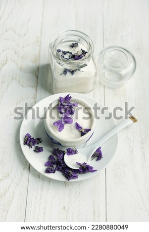 Sugar with fresh violet flowers in plate and glass jar on table close up. Edible seasonal flowers used in cooking and confectionery. Preparation of homemade healing syrup from violet flowers and sugar