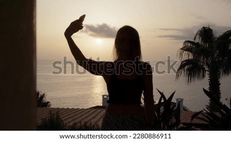 silhouette of girl in black top takes selfie on her phone against background of sunset or dawn on balcony of her room hotel. young woman takes pictures of herself on smartphone.