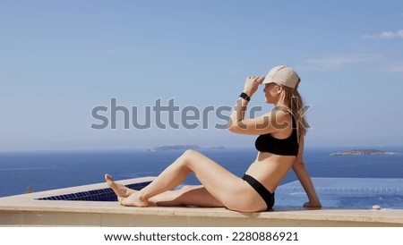 slender blonde in black swimsuit and beige cap sit on edge of pool and sunbathe, enjoys. young woman in black bikini is relaxing in hotel in relaxation area. Girl on journey on lazy vacation.