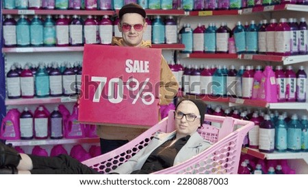 Fashionable modern man and woman looking at camera in fancy shop with 70 sale sign. rocker girl in hat sits in pink cart, guy in cap hold red sign to attract audience. discount in chemicals store.
